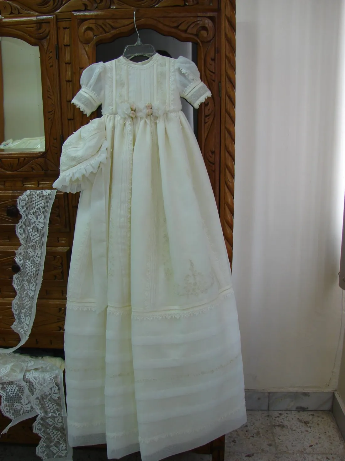 Ropón largo marfil colgado, con cofia, lazos de flores en la cintura y falda de alforzas; a un lado, tira de encaje.